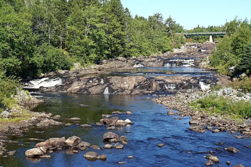 Shallow river rapids running over wide granite ledges in Sheet Harbour, Nova Scotia, with forest on both banks and a bridge in the distance