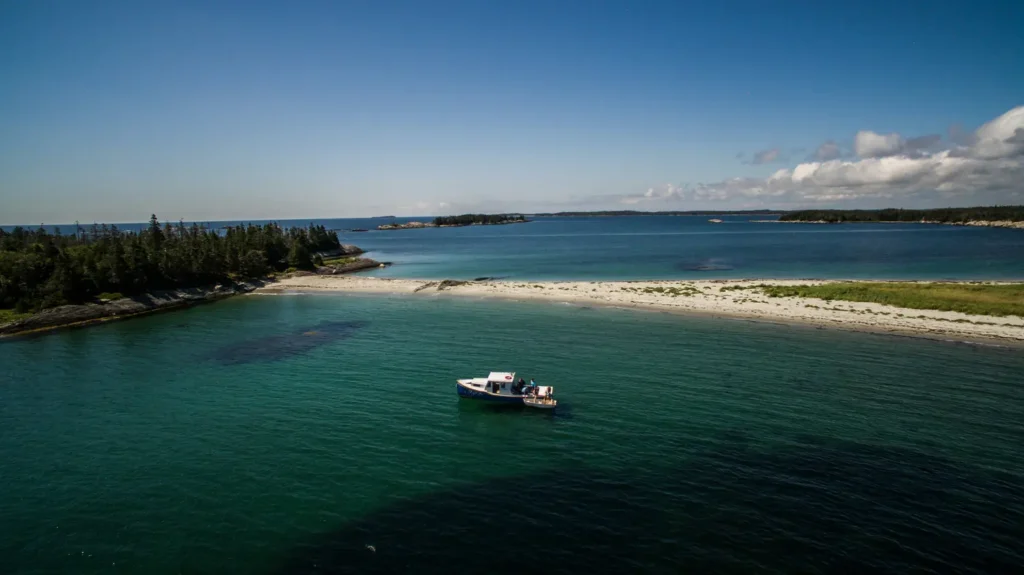 Small boat anchored in clear turquoise water beside a sandy spit and spruce-covered island in the 100 Wild Islands on Nova Scotia's Eastern Shore