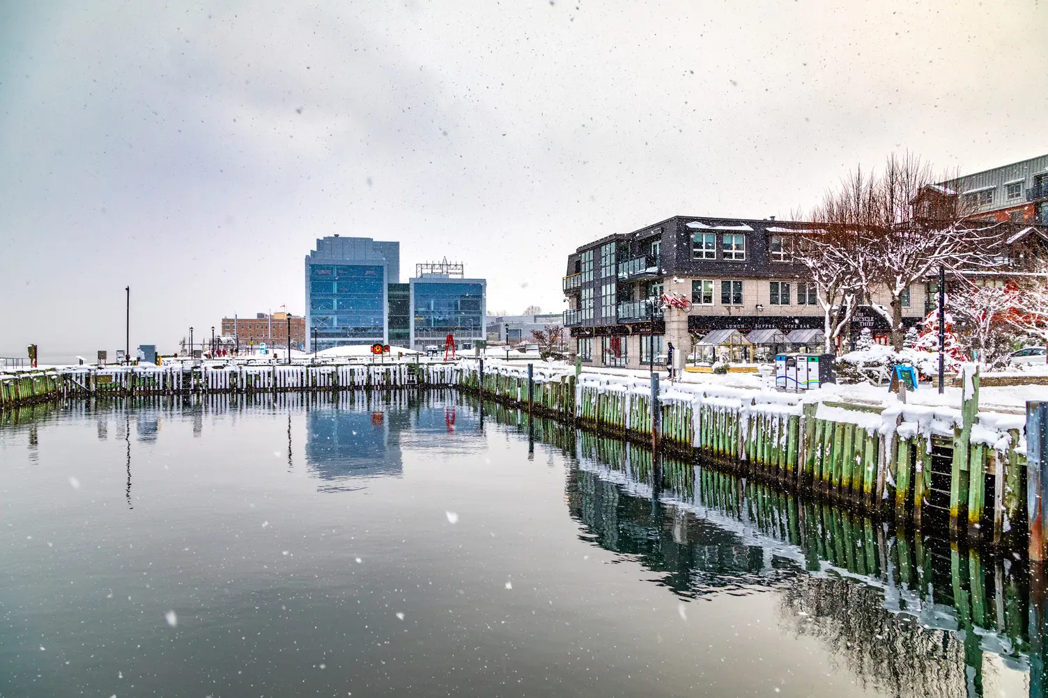 Light snow falls over the Halifax Waterfront, with a dusting of snow on the wharf and buildings reflected in the calm harbour water.