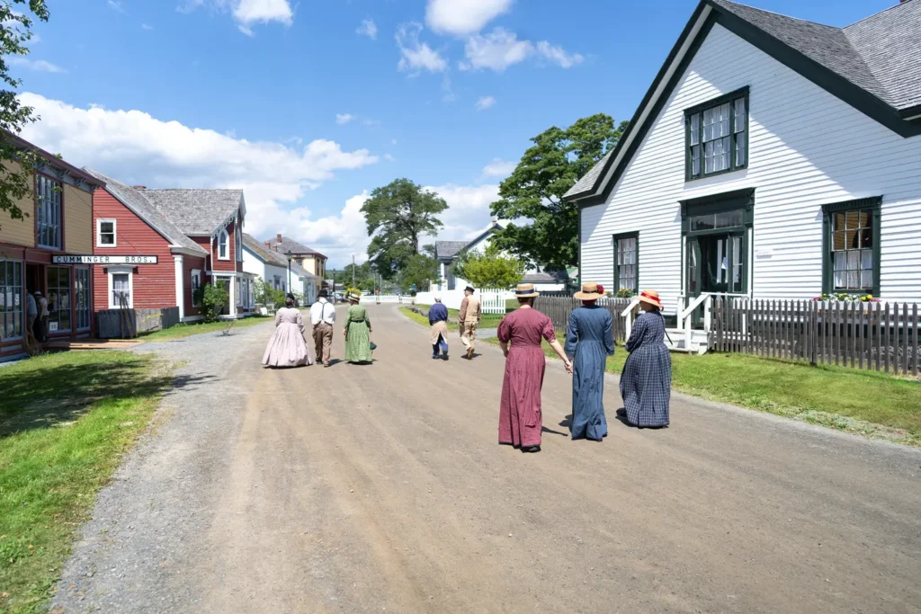 People in 19th-century-style dress walking down a gravel street lined with historic wooden buildings at Sherbrooke Village on a sunny summer day
