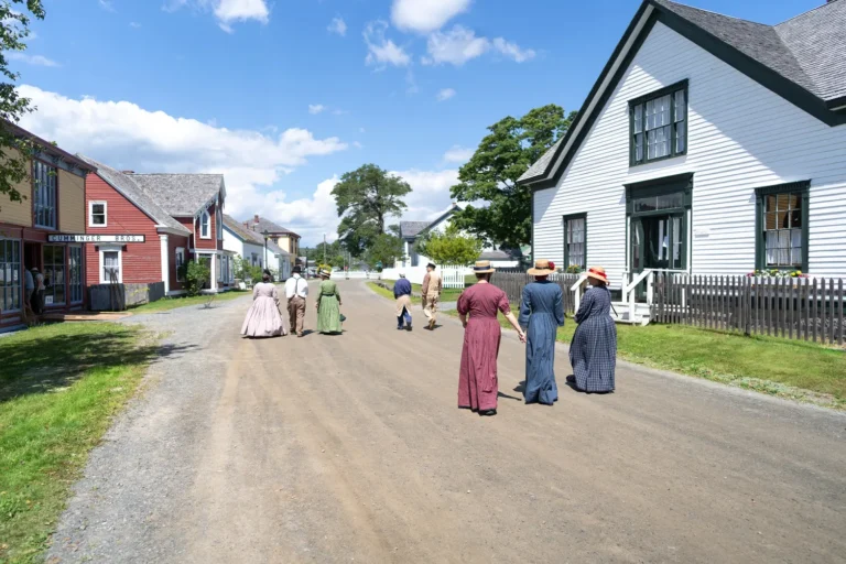 People in 19th-century-style dress walking down a gravel street lined with historic wooden buildings at Sherbrooke Village on a sunny summer day